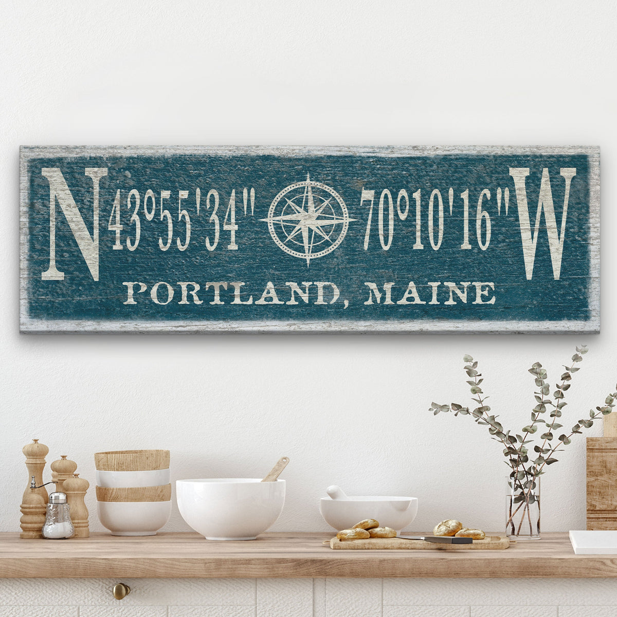 Personalized Compass Rose Canvas displaying GPS coordinates flanked by large N and W letters with a compass rose illustration at center and "Portland, Maine" at the bottom, in cream lettering on a distressed teal painted wood background, mounted above a kitchen shelf