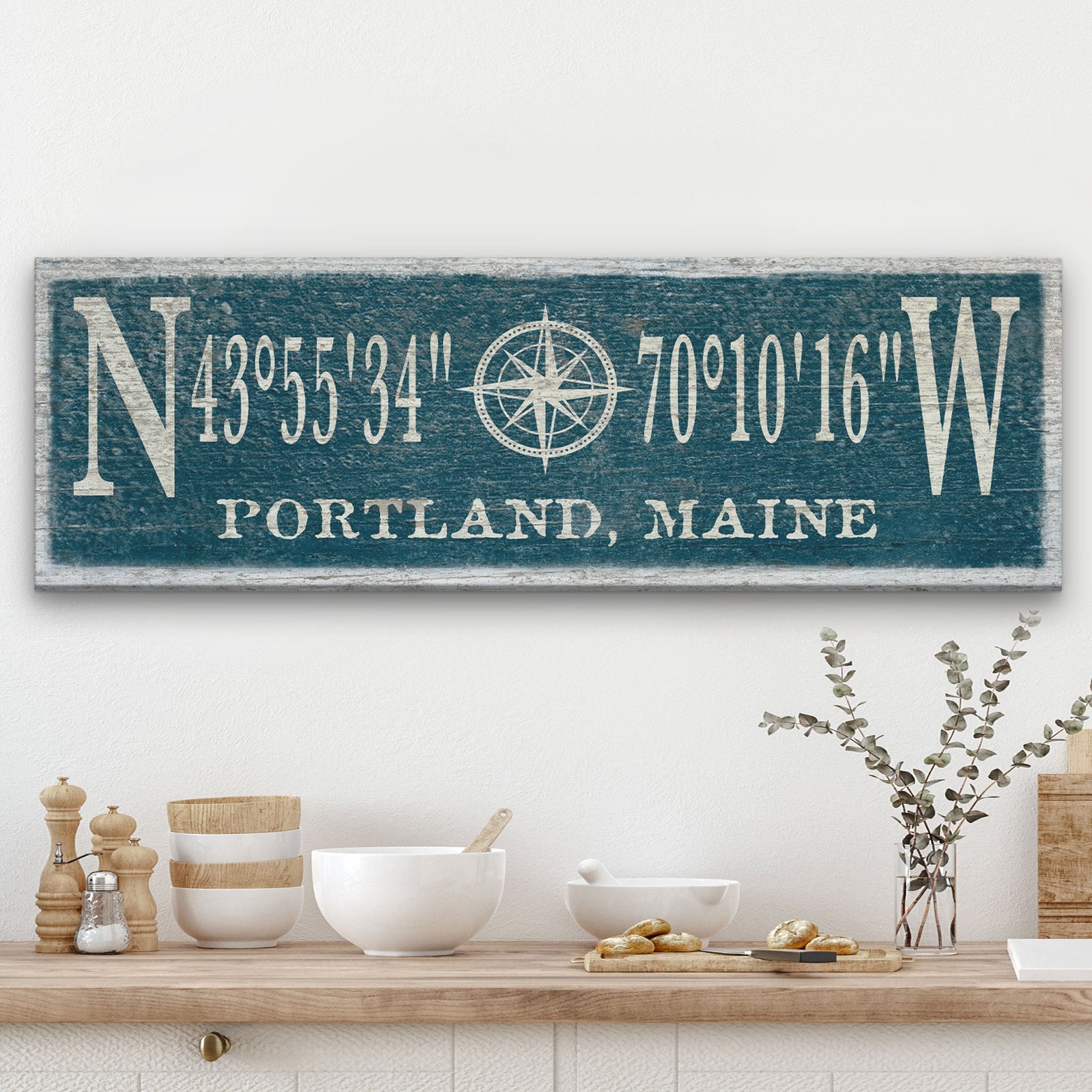 Personalized Compass Rose Canvas displaying GPS coordinates flanked by large N and W letters with a compass rose illustration at center and "Portland, Maine" at the bottom, in cream lettering on a distressed teal painted wood background, mounted above a kitchen shelf