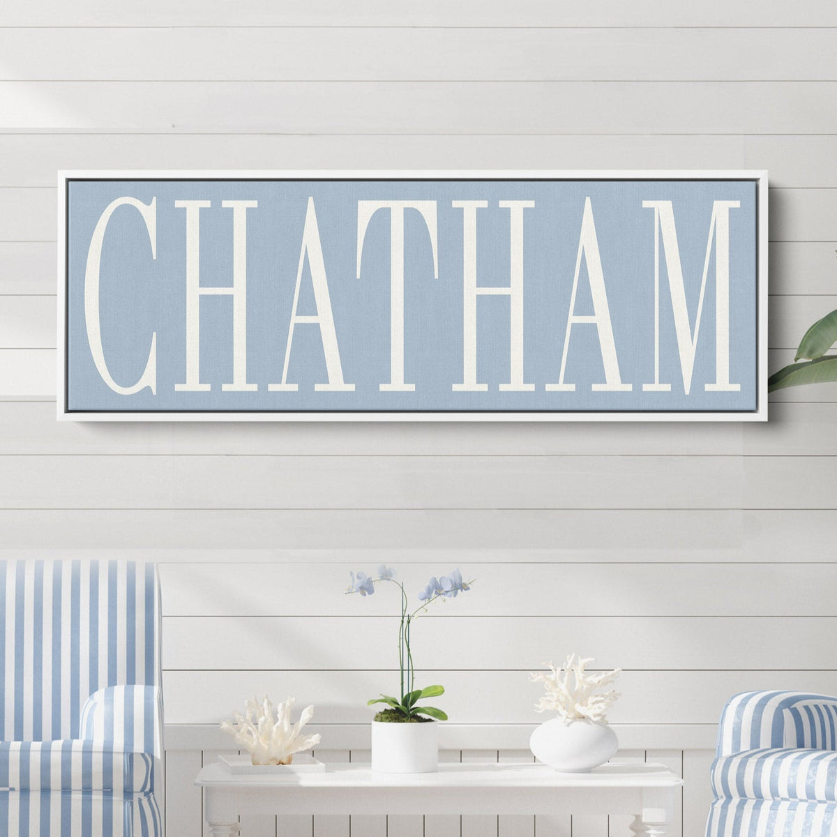 Shoreline Name Canvas showing "CHATHAM" in large white serif letters on a light blue background, displayed in a white floating frame on a shiplap wall in a coastal living room | Willow &amp; Hive