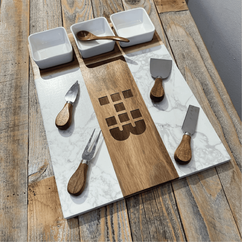 Charcuterie board set displayed on wood table with three white marble bowls, wooden spreaders and fork with wood handles, and center board engraved with sample logo design.