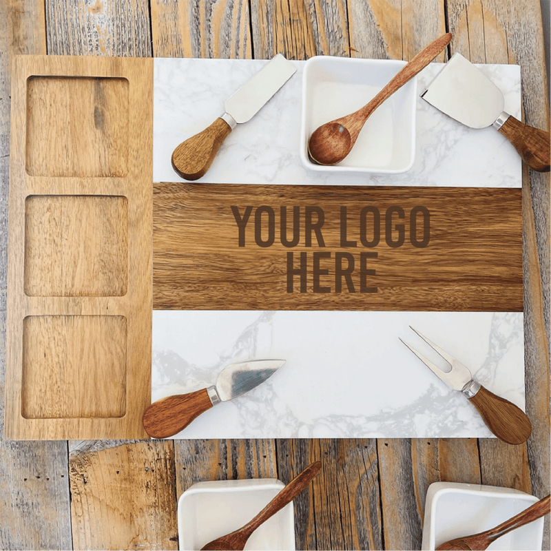 Overhead view of marble and wood charcuterie board with sample logo text, surrounded by wooden spreaders, small bowls, and serving utensils arranged on wooden surface