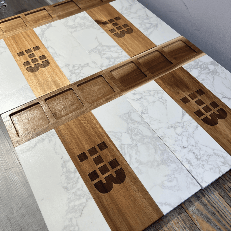 Multiple custom logo charcuterie boards arranged in flat lay, showing engraved monogram designs on acacia wood strips alternating with white marble tiles and matching marble bowls