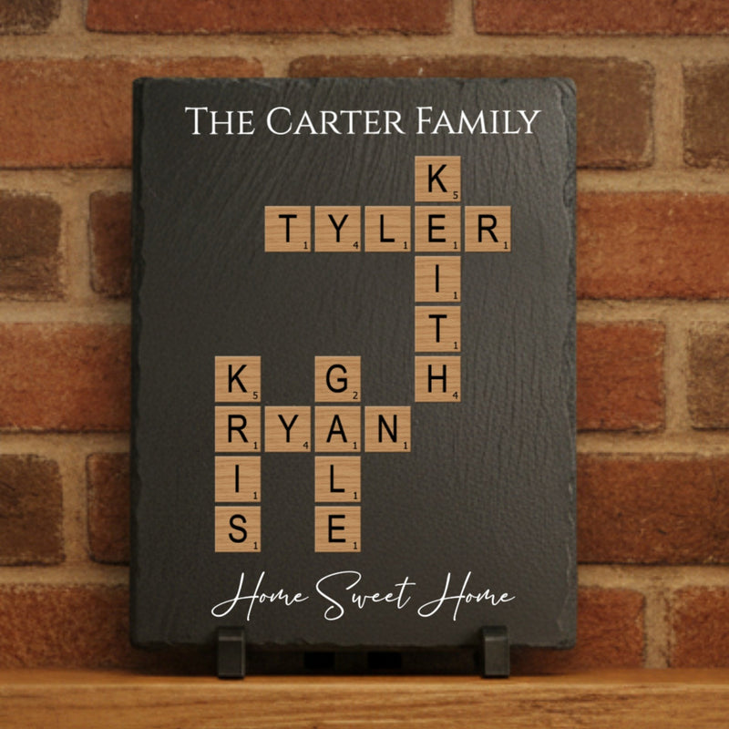 Dark slate board with sample family names "TYLER," "RYAN," and "KITE" arranged in crossword pattern using Scrabble-style tiles, with "The CARTER FAMILY" engraved at top and "Home Sweet Home" at bottom