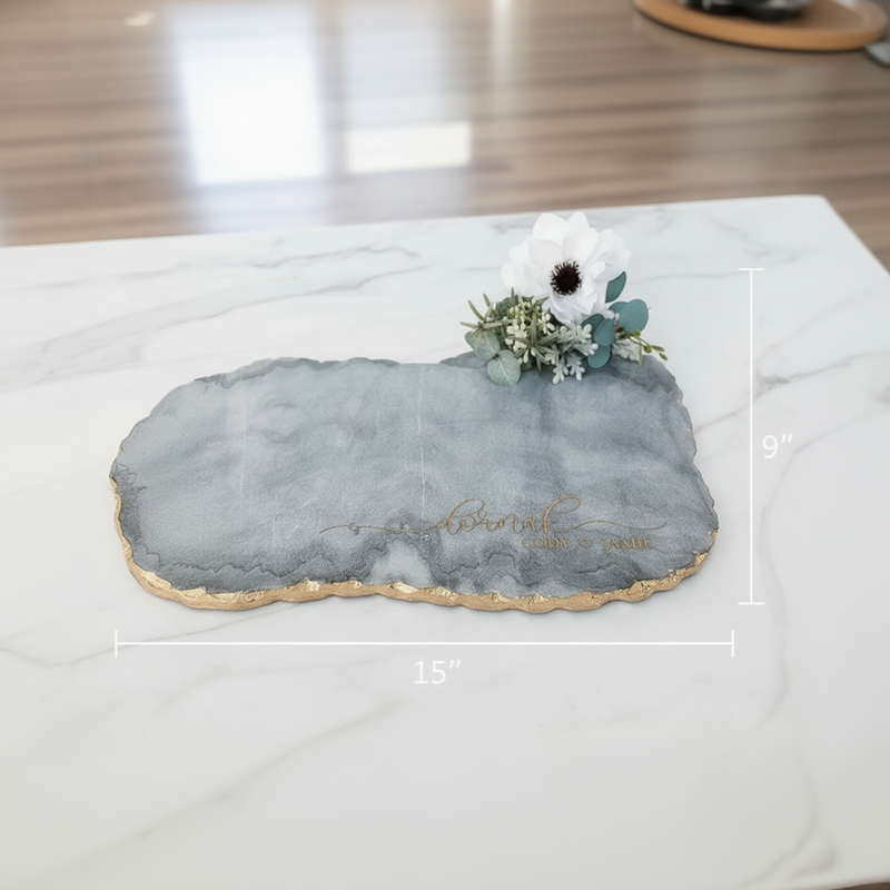 Gray and white marble serving board with gold-leafed edge, displaying sample engraving and topped with white daisy and eucalyptus floral arrangement, shown with dimensions 15" by 9