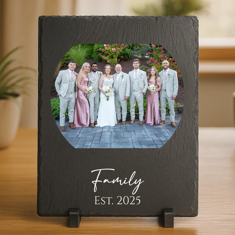Dark slate frame displaying a wedding photo of nine people in formal attire with flowers, engraved with sample text "Family" and "Est. 2025" below the image