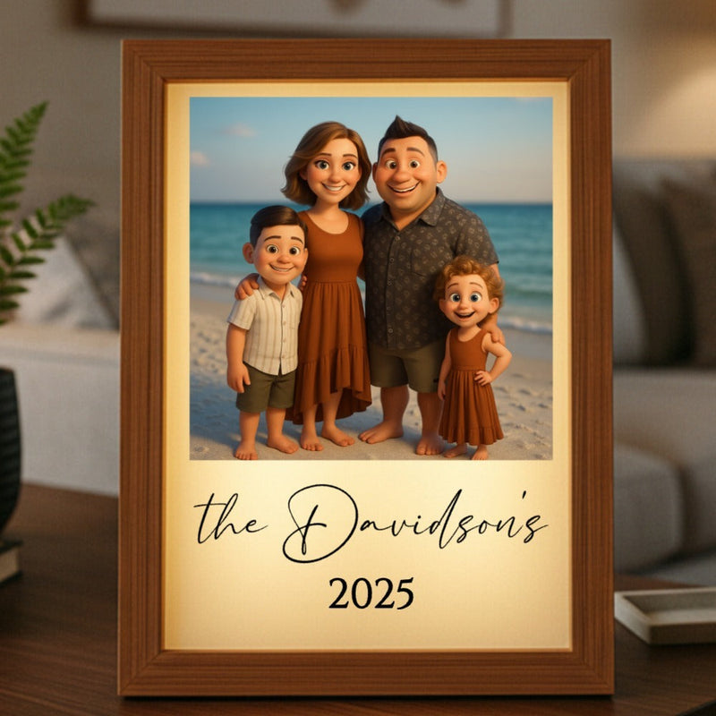 Framed light box displaying animated family portrait on beach with sample text "the Davidsons" and "2025" on cream-colored mat, wood frame displayed on wooden surface