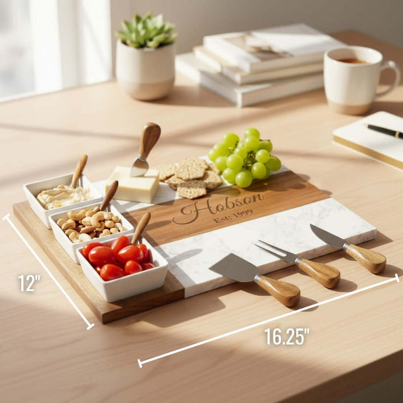 Wooden charcuterie board with sample engraving "Hobson Est. 1999," three white ceramic bowls with spreaders holding nuts and tomatoes, cheese, crackers, green grapes, and cheese knives, displayed on desk with dimensions 16.25" by 12