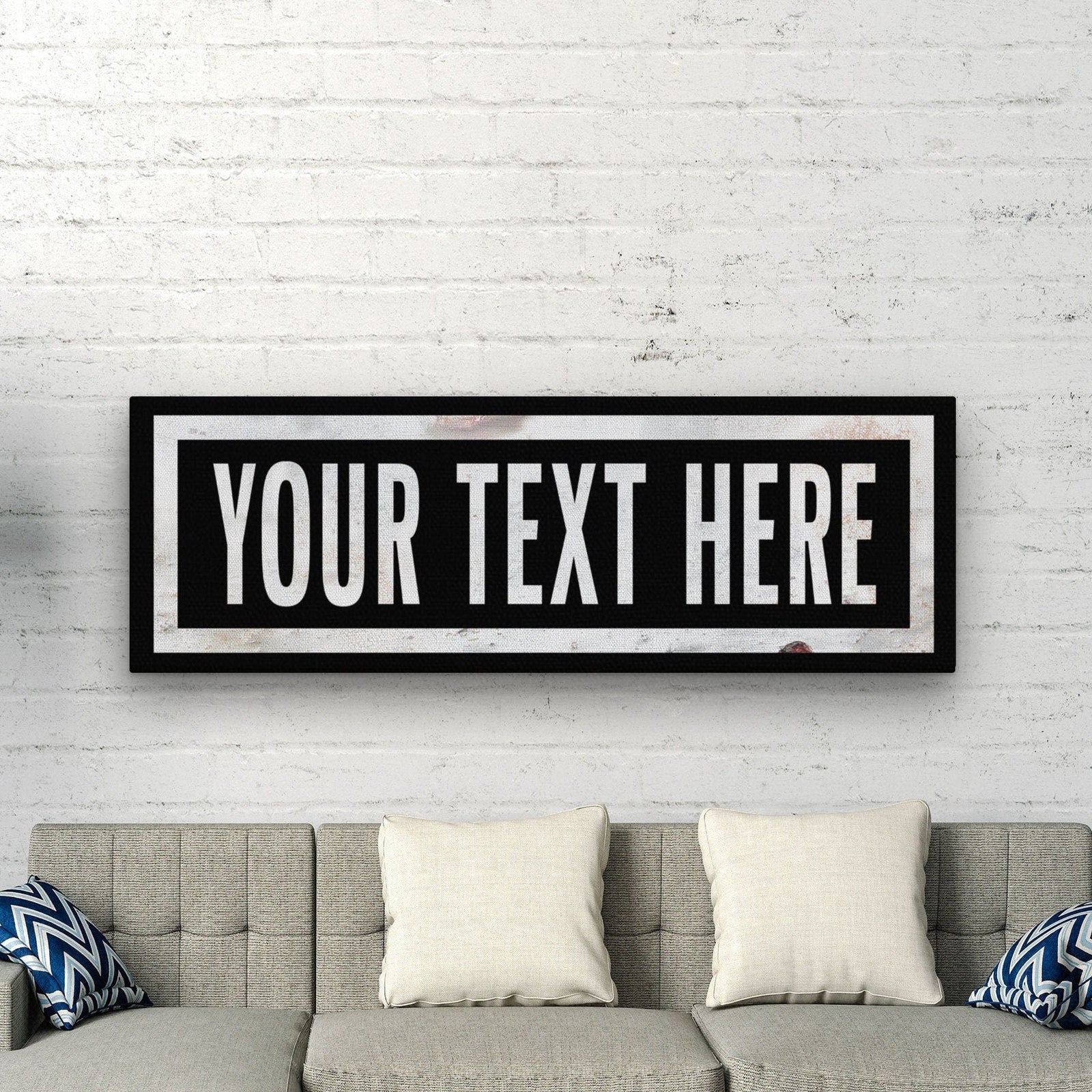 Personalized marquee canvas hanging above a gray upholstered sofa with cream pillows, displaying sample text "YOUR TEXT HERE" in white letters on black background with double border frame