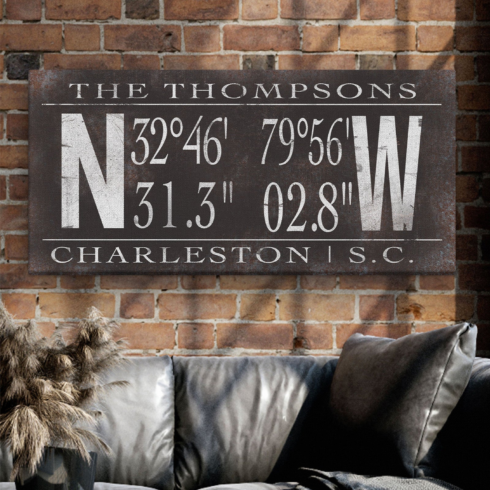 Dark charcoal canvas with sample family name "THE THOMPSONS" and GPS coordinates 32°46' 79°56' with location "CHARLESTON | S.C." displayed on brick wall above gray couch