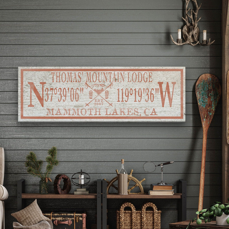 Personalized canvas ski lodge sign with sample name "Thomas Mountain Lodge," coordinates, and location "Mammoth Lakes, CA" in rust and cream colors, displayed on a dark shiplap wall above cabin decor.