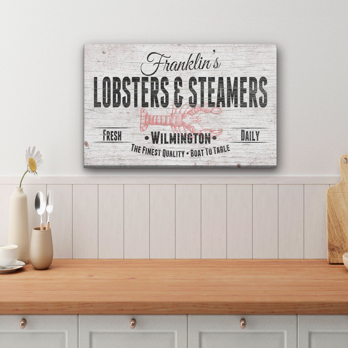 Personalized canvas sign with sample name "Franklin's" and location "Wilmington" displayed on kitchen wall above wooden counter with white shiplap backsplash