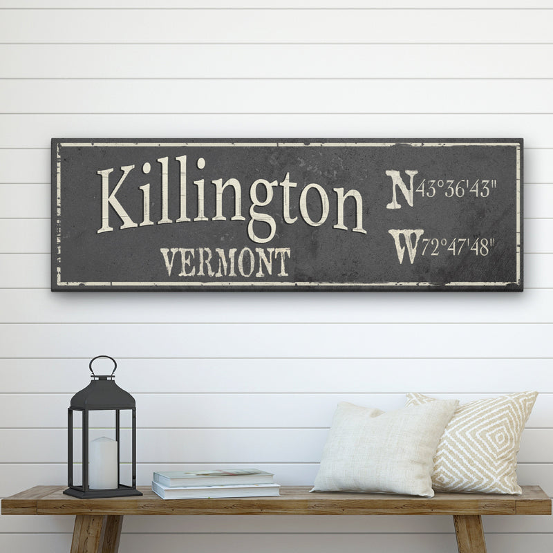 Dark gray canvas sign displaying "Killington" with sample coordinates "N43°36'43" W72°47'48"" and "Vermont" in cream lettering, mounted above a wooden shelf.