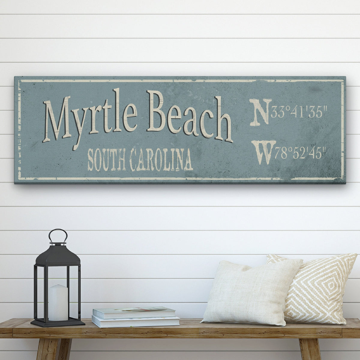 Personalized Hometown Canvas displaying "Myrtle Beach" in large cream serif lettering with "South Carolina" in caps below, alongside stacked N and W GPS coordinates on the right side, all on a distressed dusty blue background with a cream painted border frame, mounted on a shiplap wall above a rustic bench with lantern