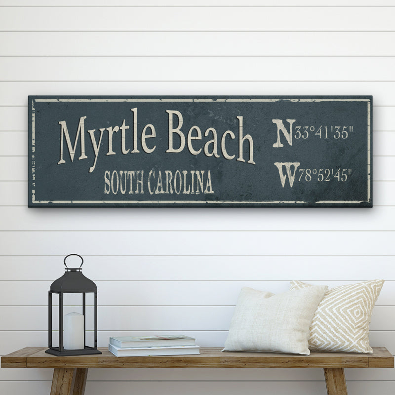 Distressed dark blue canvas sign displaying "Myrtle Beach" with "South Carolina" and GPS coordinates, hung above wooden bench with lantern and pillows