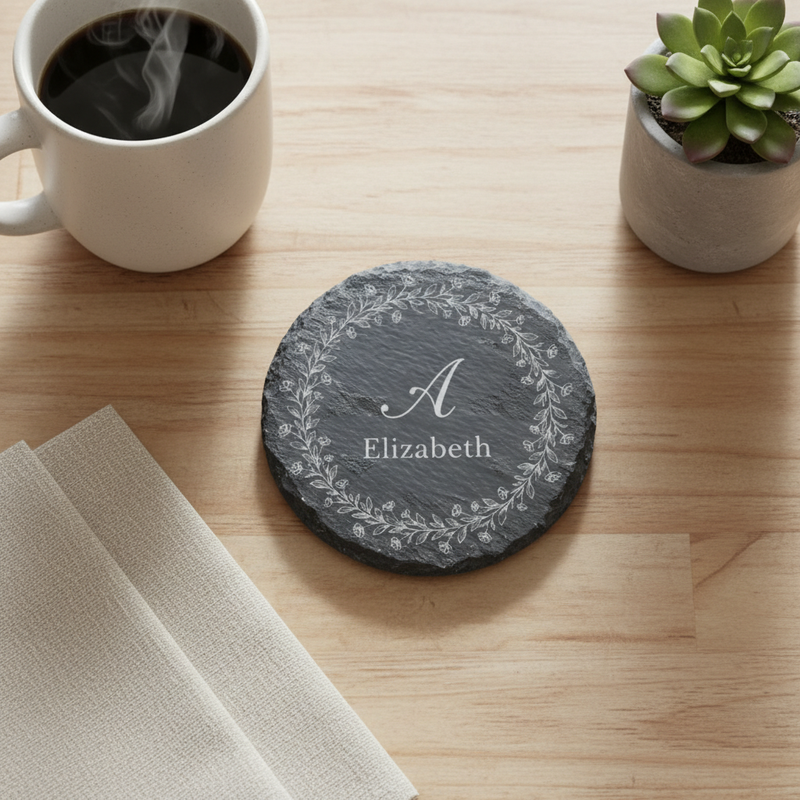 Slate coaster engraved with sample initial "A" and name "Elizabeth" surrounded by floral wreath, displayed on wooden surface with coffee mug and succulent plant