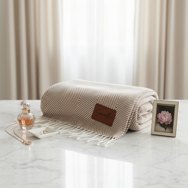 Beige herringbone throw blanket with fringe edges, displayed on marble surface alongside perfume bottle, jewelry dish, and framed peony photo, styled with sheer curtains in background