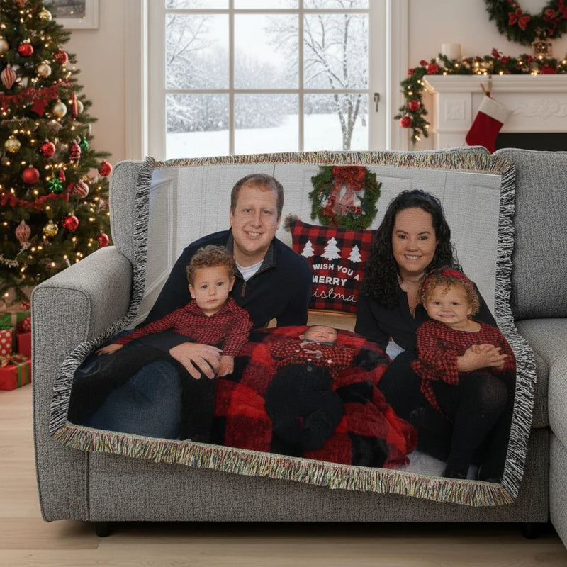 Family of four sitting on a gray sofa draped with a personalized woven photo throw blanket featuring their holiday portrait, with colorful fringe trim around edges.