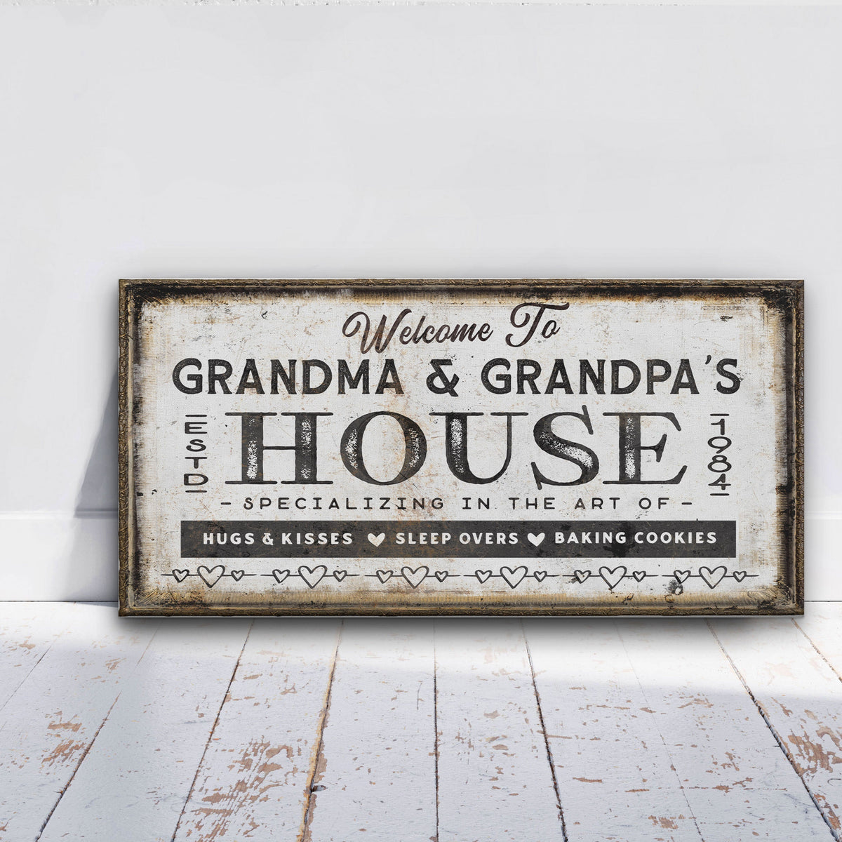 Grandparents' House Canvas displaying "Welcome To Grandma &amp; Grandpa's House Est. 1984" with "Specializing in the Art of Hugs &amp; Kisses · Sleep Overs · Baking Cookies" in bold black distressed lettering on an aged cream background with a dark rustic floating frame