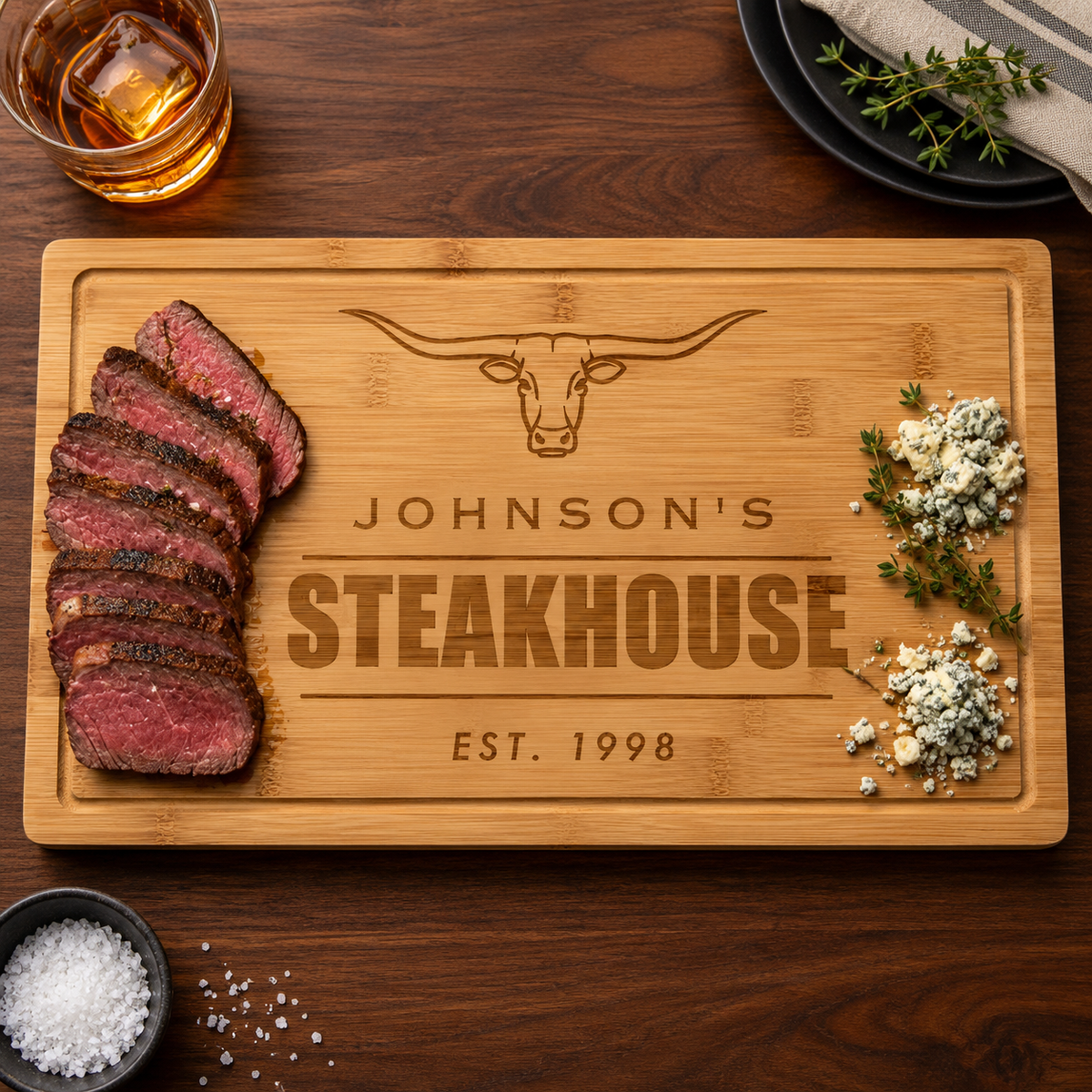 Wooden cutting board engraved with longhorn design and text reading "Johnson's Steakhouse Est. 1998," styled with sliced steak and fresh herbs