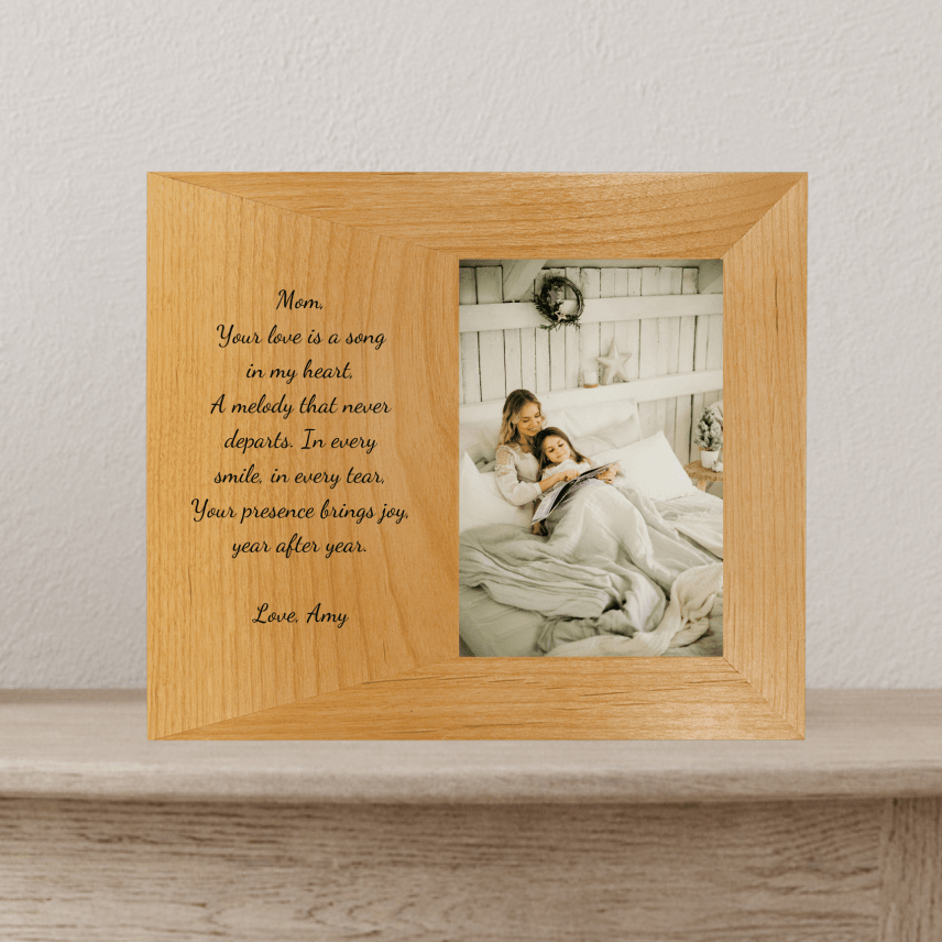 Light wood picture frame with engraved sample text reading "Mom, Your love is a song in my heart..." and "Love, Amy" on left side, displaying photo of mother and child reading in bed