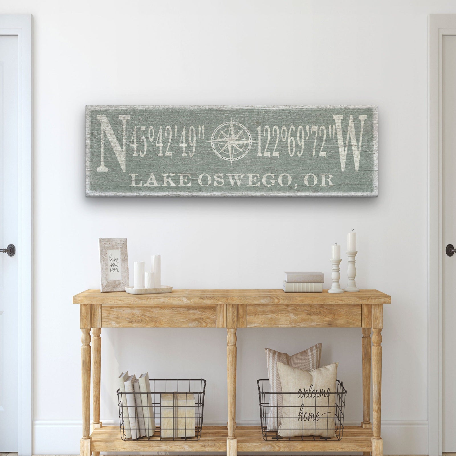Sage green canvas sign hanging above wooden console table, engraved with sample coordinates and "Lake Oswego, OR" with centered compass rose design.