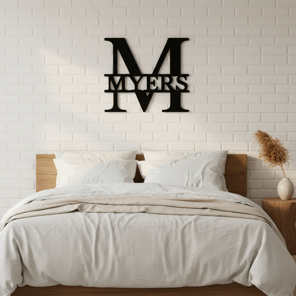 Black metal monogram wall sign with sample name "MYERS" displayed above a bed with white bedding and wooden nightstands against white brick.