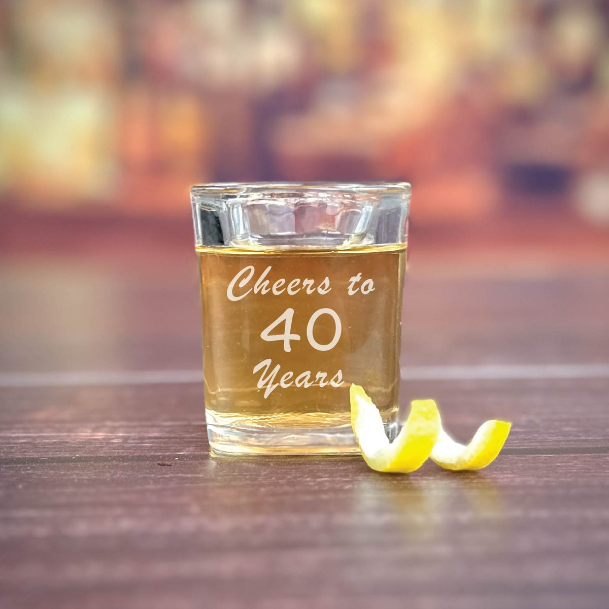 Shot glass filled with amber liquid, engraved with sample text "Cheers to 40 years" in white script, lemon twist beside it on wooden surface