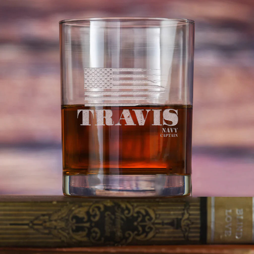 Rocks glass filled with whiskey, engraved with tattered American flag and sample name "TRAVIS" with "NAVY CAPTAIN" below, positioned on wooden surface against blurred background