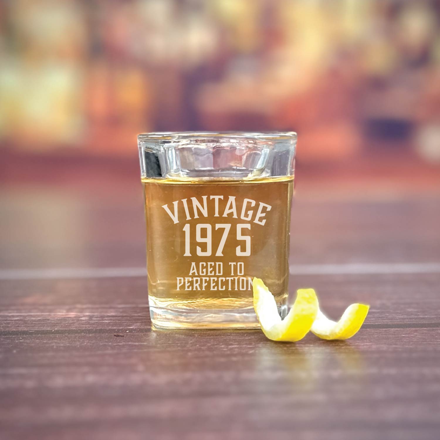 Clear shot glass filled with amber liquid, engraved with sample text "Vintage 1975 Aged to Perfection," with lemon twist beside it on wooden surface
