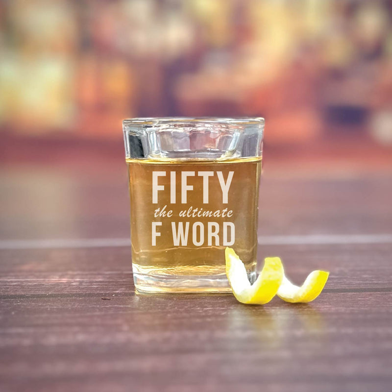 Shot glass filled with amber liquid, engraved with "FIFTY the ultimate F WORD" in white text, lemon twist beside it on wooden surface