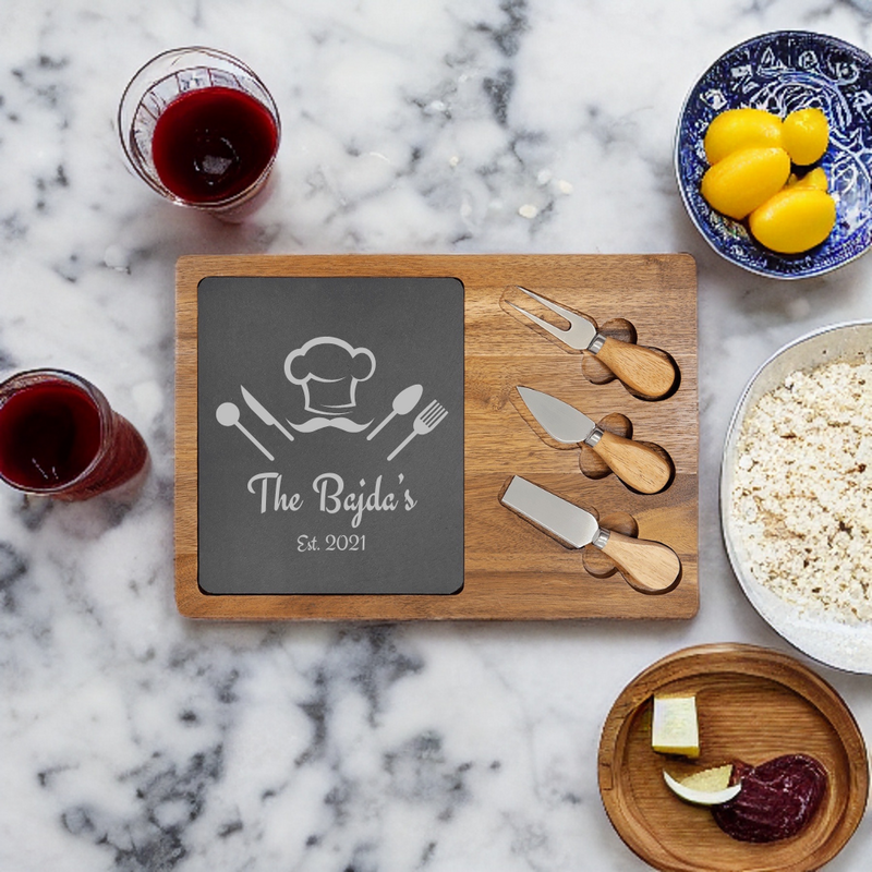 Wooden charcuterie board with slate inlay engraved with sample text "The Bejda's Est. 2021" and chef hat icon, surrounded by three spreader knives with wooden handles, displayed with wine glasses, lemons, and food items on marble surface.