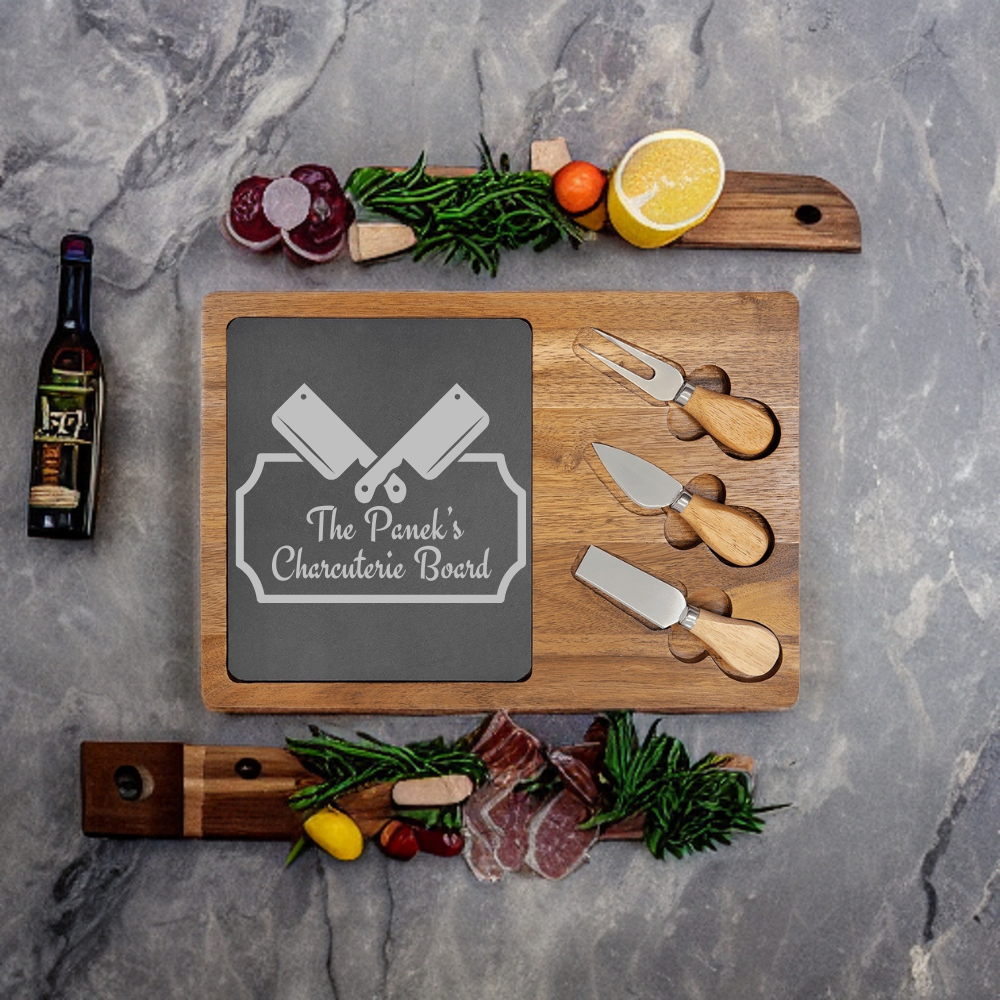 Wooden charcuterie board with slate inlay engraved with sample text "The Panek's Charcuterie Board," displayed with three cheese knives and fresh ingredients including meats, greens, lemon, and wine.