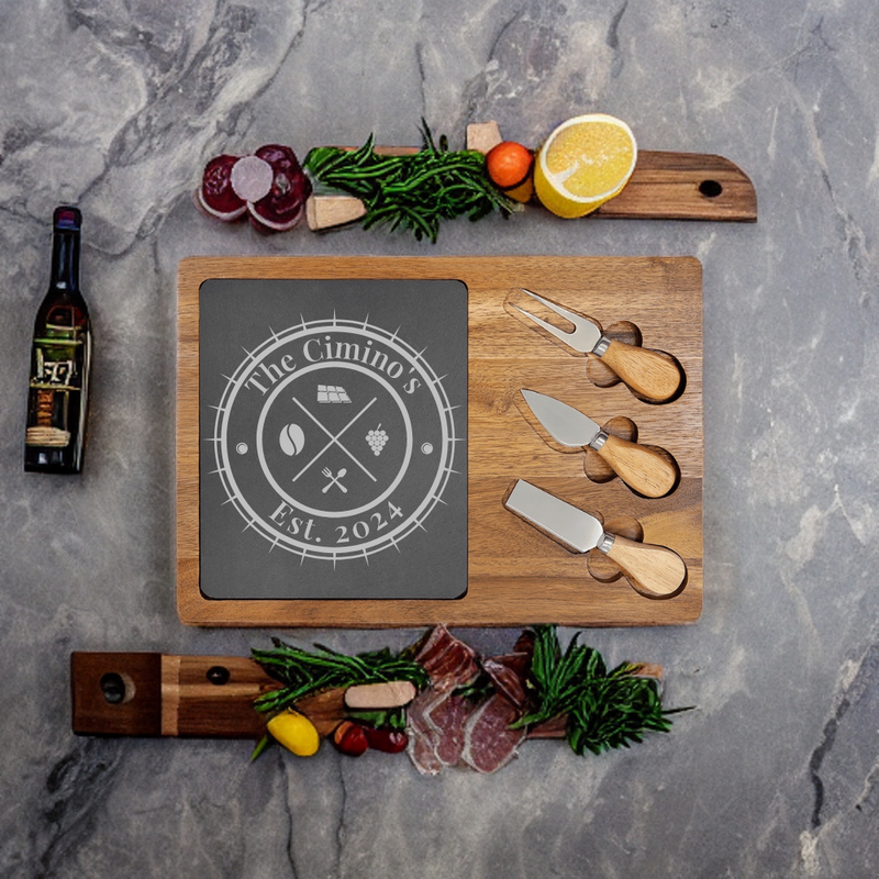 Wooden charcuterie board with slate center engraved with sample family name "The Cimino's" and "Est. 2024", accompanied by three cheese knives with wooden handles and fresh ingredients arranged on gray marble surface.