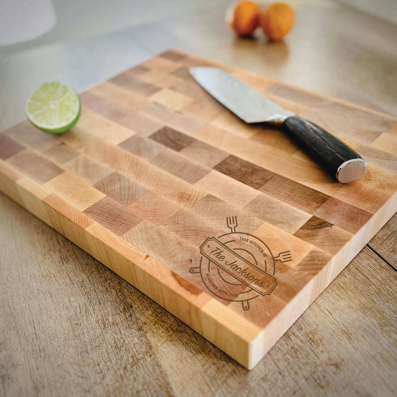 Maple butcher block with checkered surface pattern and engraved kitchen design reading "This Kitchen" with fork and spatula motifs, styled with lime half and chef's knife on wooden table