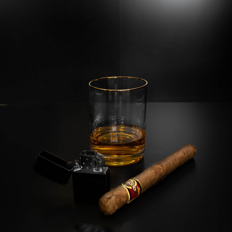 Gold-rimmed whiskey glass filled with amber liquid, displayed alongside a cigar and lighter on dark surface