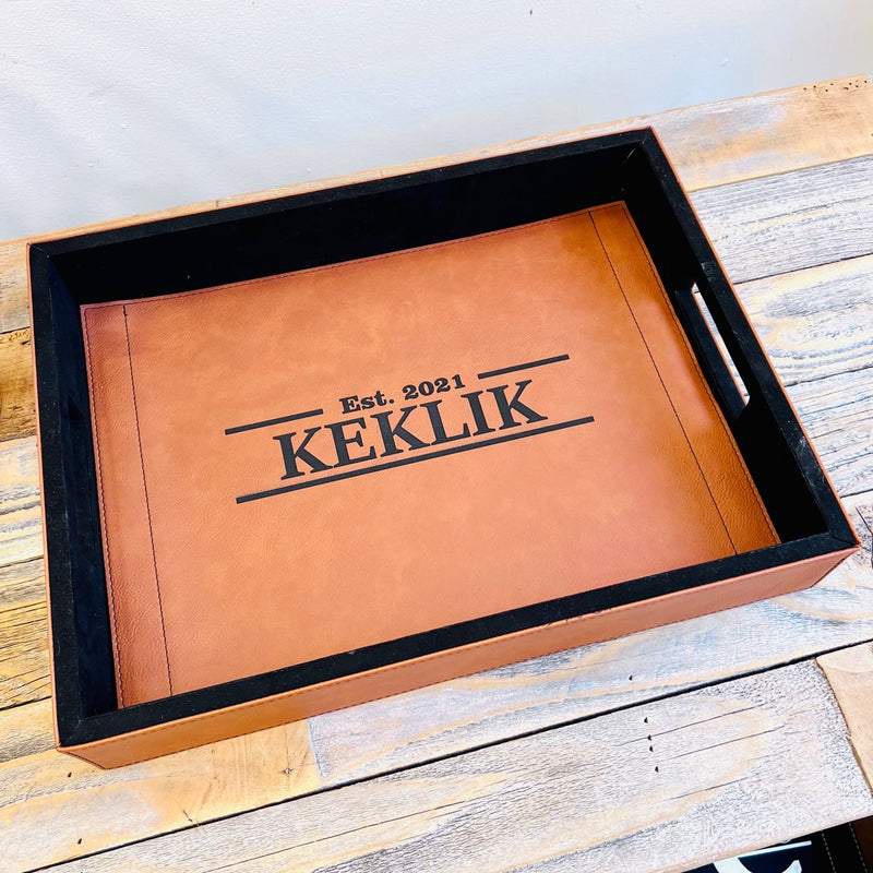 Tan leather serving tray personalized with sample engraving "Est. 2021 KEKLIK" displayed in black wooden box on whitewashed wood surface
