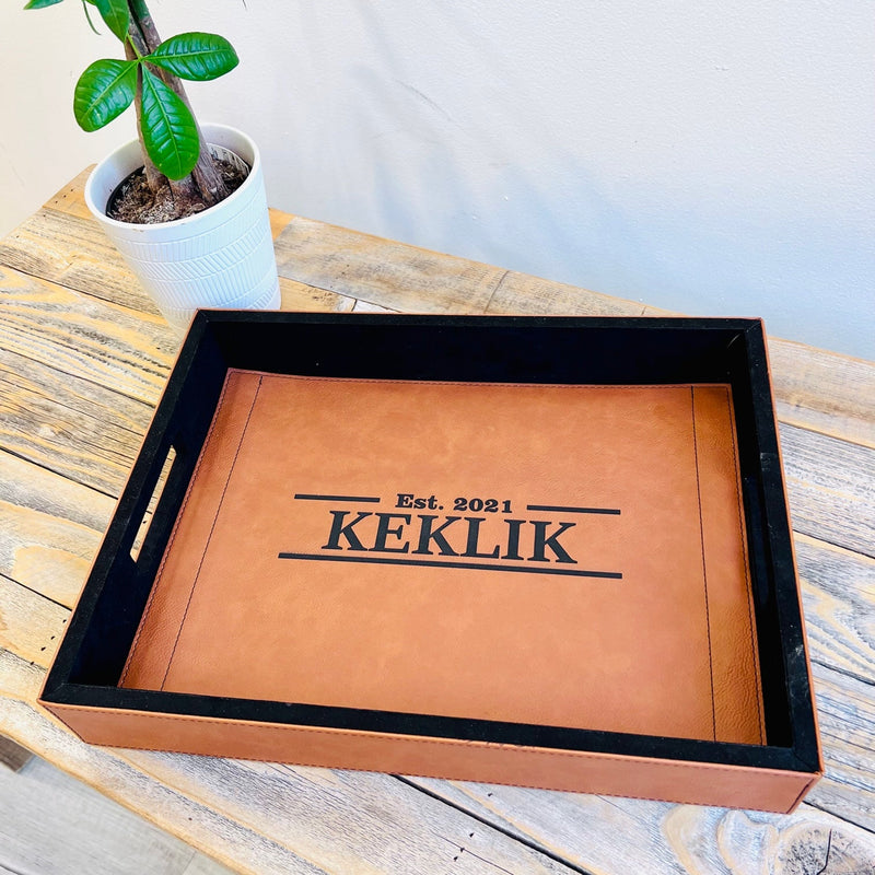 Tan leather serving tray with black interior, laser-engraved with sample text "Est. 2021 KEKLIK" and decorative lines, displayed on wooden table with potted plant.
