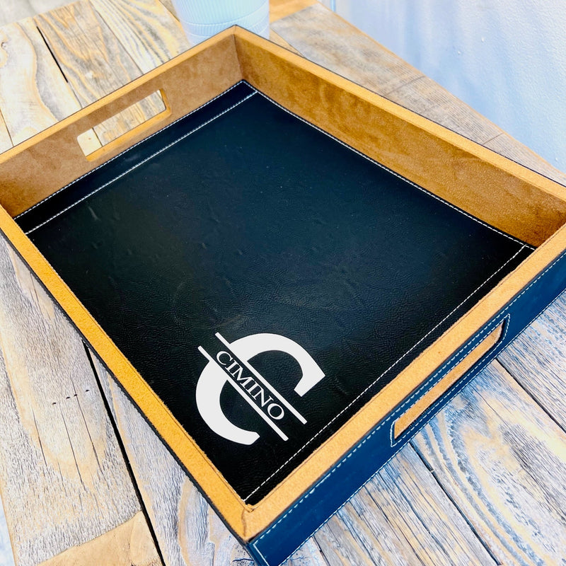 Black leather serving tray with wood frame, displaying engraved logo on interior surface, viewed from above on wooden background