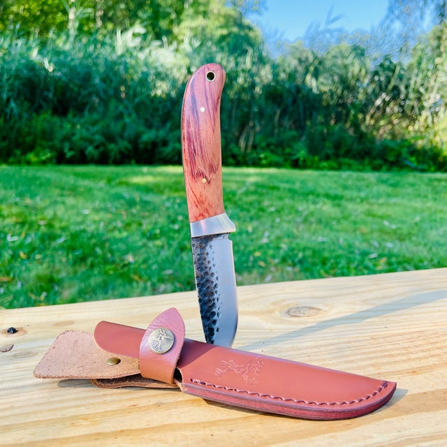 Fixed blade hunting knife with wooden handle and dark blade displayed on wooden surface next to brown leather sheath with metal emblem