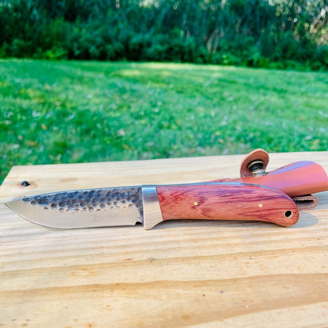 Fixed blade knife with drop point blade and wood handle resting on light wooden surface, with green grass and shrubs blurred in background