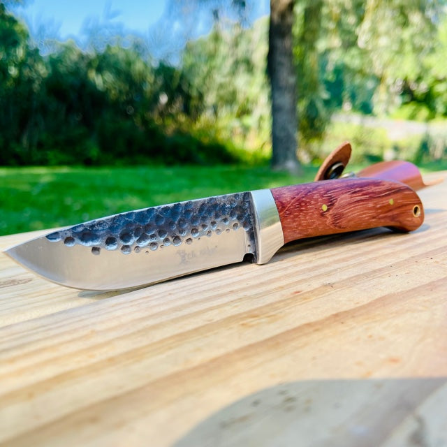 Fixed blade knife with hammered blue steel blade and wood handle resting on light wooden surface outdoors, with green garden background