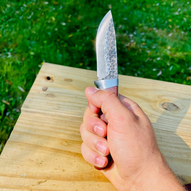 Hand holding fixed blade knife with textured handle over wooden surface, green grass background
