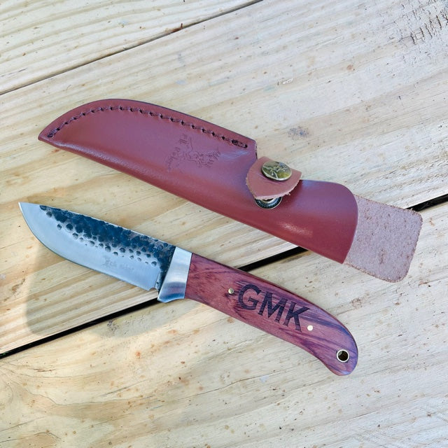 Fixed blade knife with hammered steel blade and burgundy Pakkawood handle engraved with sample initials "GMK", displayed with matching leather sheath on wooden surface