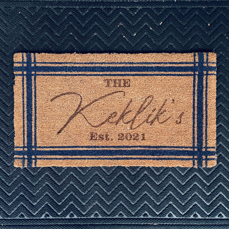 Cork welcome mat with navy blue border and sample engraving showing "Est. 2021" and decorative script, displayed on textured dark fabric background