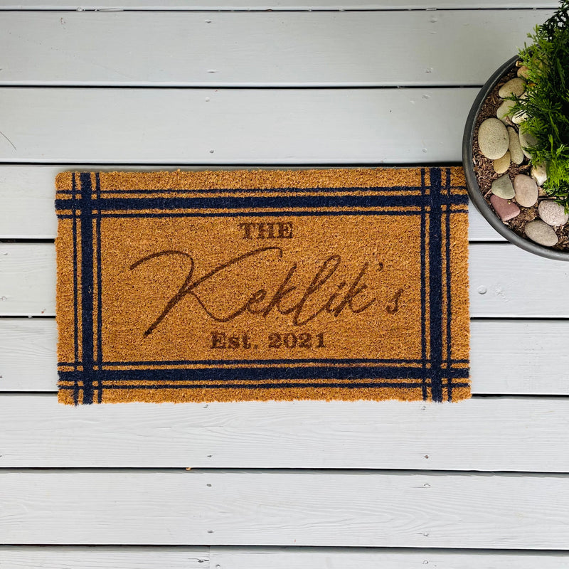 Cork welcome mat with navy blue border detail, personalized with sample name "The Rileys" and "EST. 2021" engraved in the center, displayed on white wooden deck