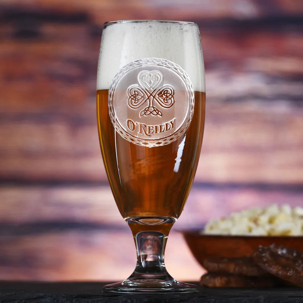 Tall beer goblet filled with amber beer and foam head, engraved with Celtic shamrock design and sample name "O'REILLY" in circular frame, set against blurred sunset background