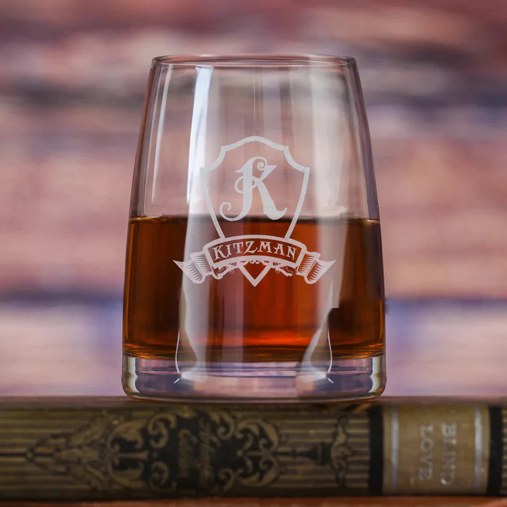 Crystal rocks glass engraved with decorative shield crest and sample name "KITZMAN" filled with whiskey, positioned against blurred sunset background