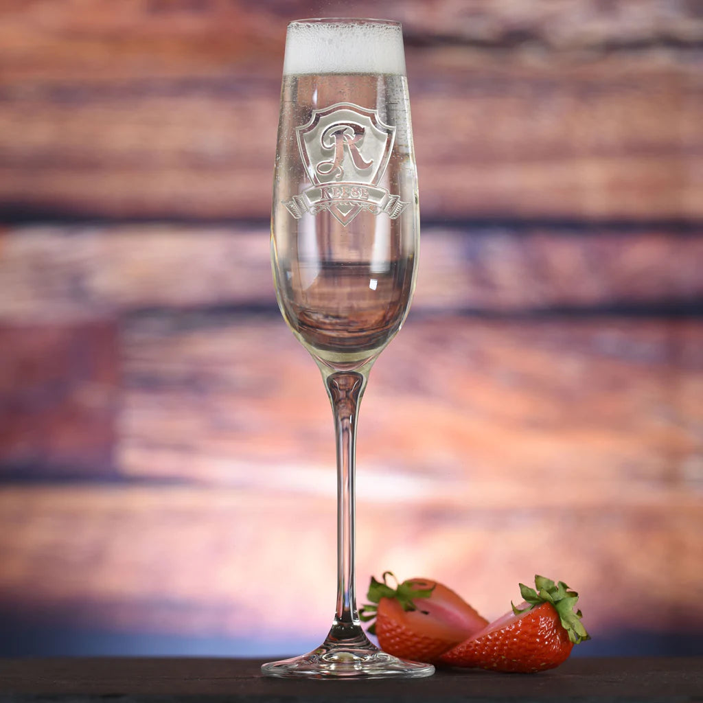 Crystal champagne flute with engraved shield and monogram letter "R" design, filled with sparkling beverage, displayed against blurred sunset background with fresh strawberries