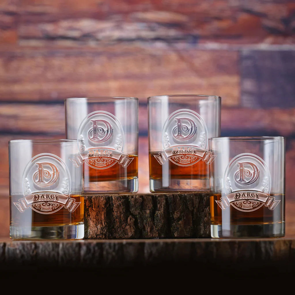 Four clear bourbon glasses with engraved "P" monogram and "Darcy" banner, filled with whiskey, arranged around a dark wood block against a brick backdrop.