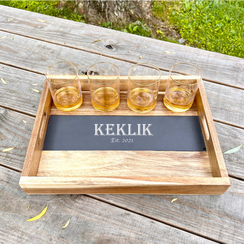 Wooden serving tray with removable slate insert engraved with sample name "KEKLIK" and "Est. 2021", holding four gold-rimmed whiskey glasses filled with amber liquid, displayed on outdoor wooden deck.